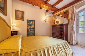 A bedroom with a bed, a dresser, a wall-mounted shelf with books and decorative items, a painting, and a window with curtains. - Villa Goffrey, Sicily (San Ciro - Ulmi - Filci)