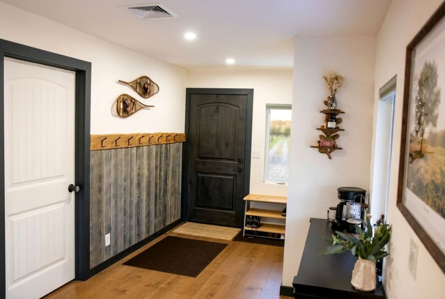 Un pasillo con suelo de madera, una puerta blanca, una puerta de madera oscura y una pequeña mesa con una planta.