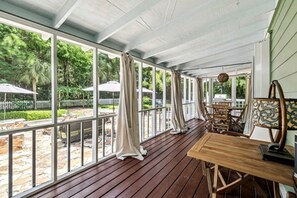 Ein überdachter Außenbereich mit Holzdeck, einem Tisch und Stühlen und Blick auf einen Hinterhof mit weißem Zaun und Grünpflanzen.