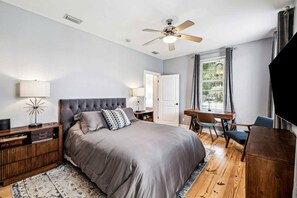 Ein Schlafzimmer mit Bett, Nachttischen, Schreibtisch, Stuhl, Deckenventilator und einem Fenster mit Vorhängen.