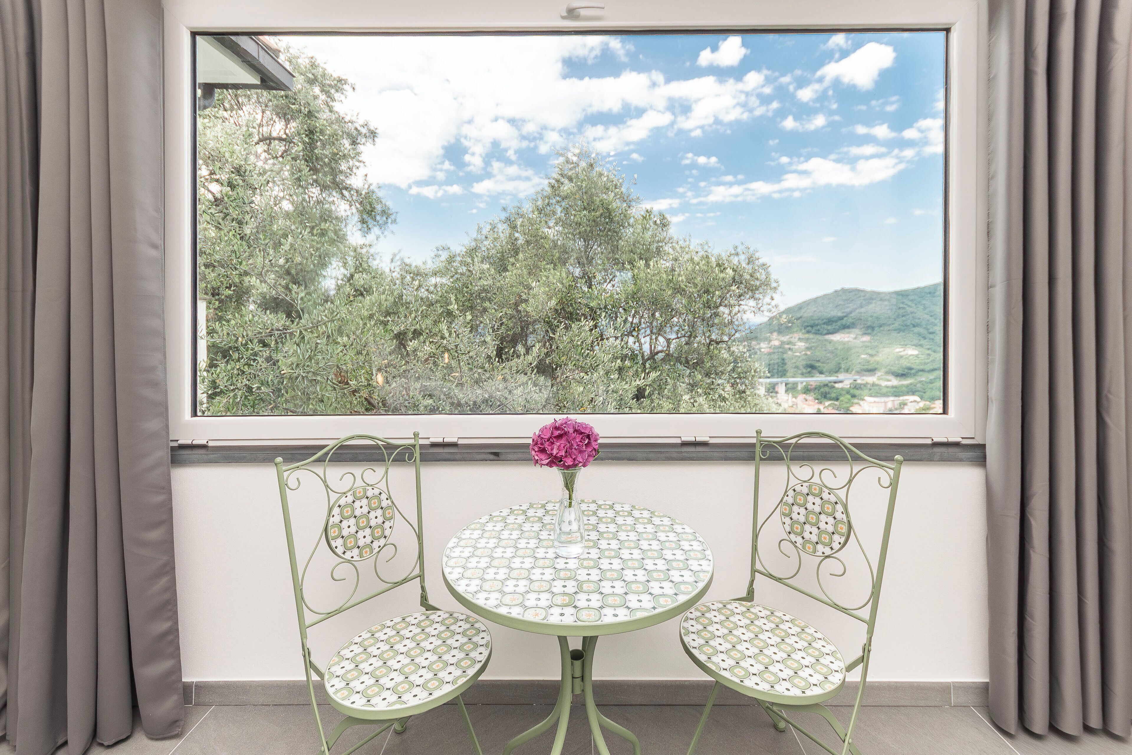 Un balcone con tavolo e due sedie, un vaso con un fiore e vista su alberi e colline.
