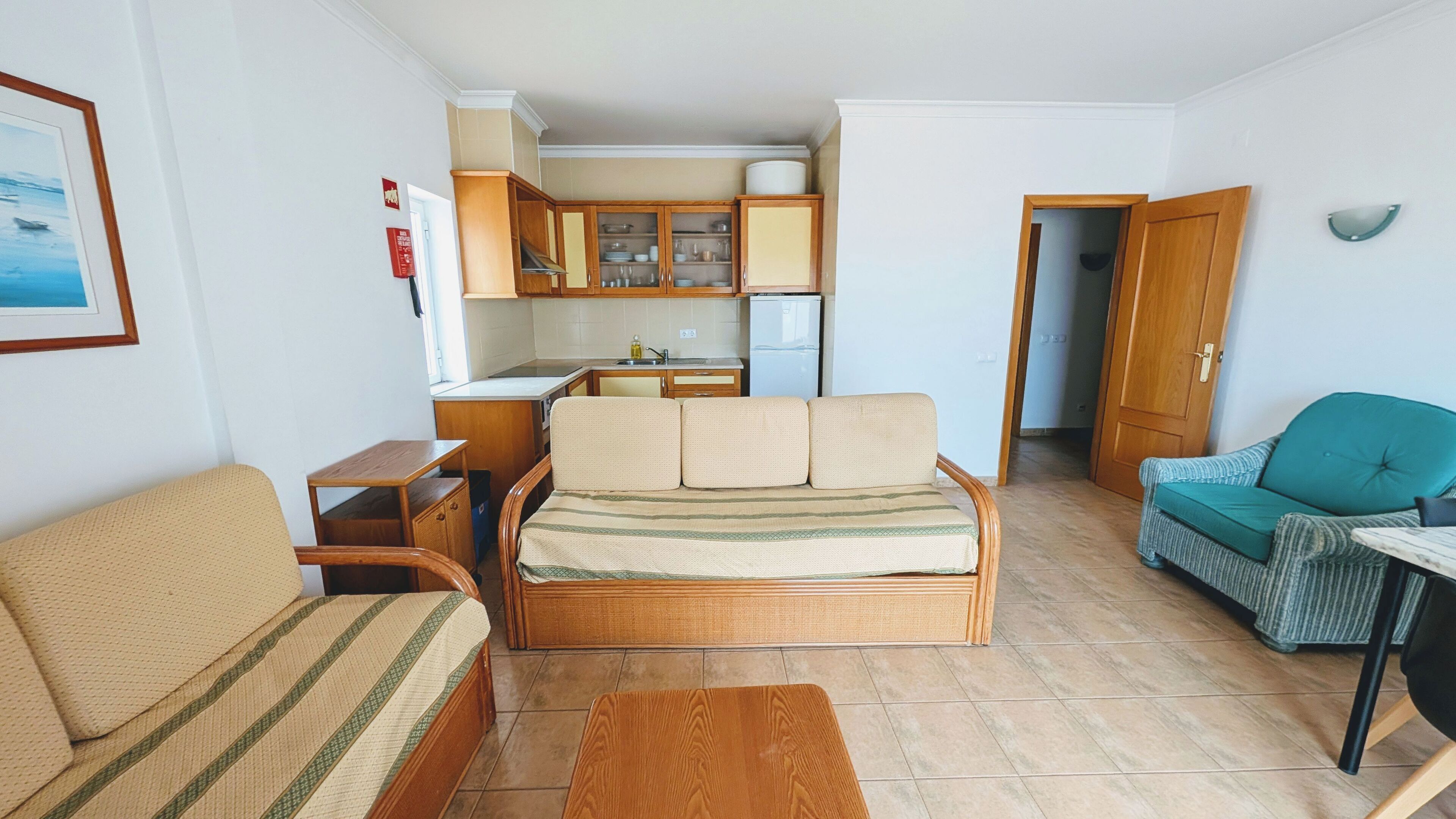 A compact living space with a sofa, a small table, a kitchenette with cabinets and a refrigerator, and a door leading to another room.