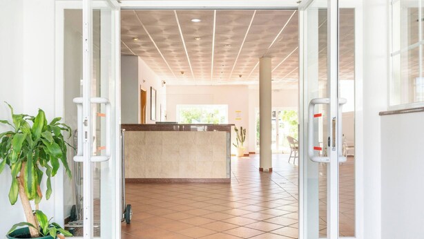 Un vestíbulo de hotel con mostrador de recepción, puertas abiertas, suelo de baldosas y plantas en macetas.