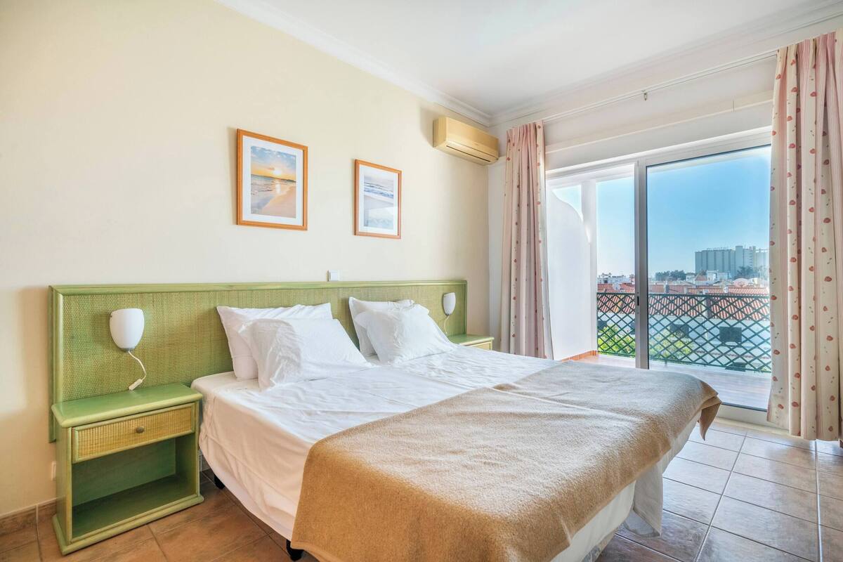 Una habitación de hotel con cama, mesita de noche, ventana con vistas y un cuadro en la pared.