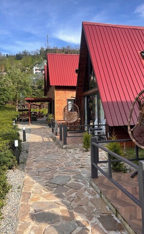 A stone pathway leading to a wooden building with a red roof, surrounded by greenery and a clear sky.