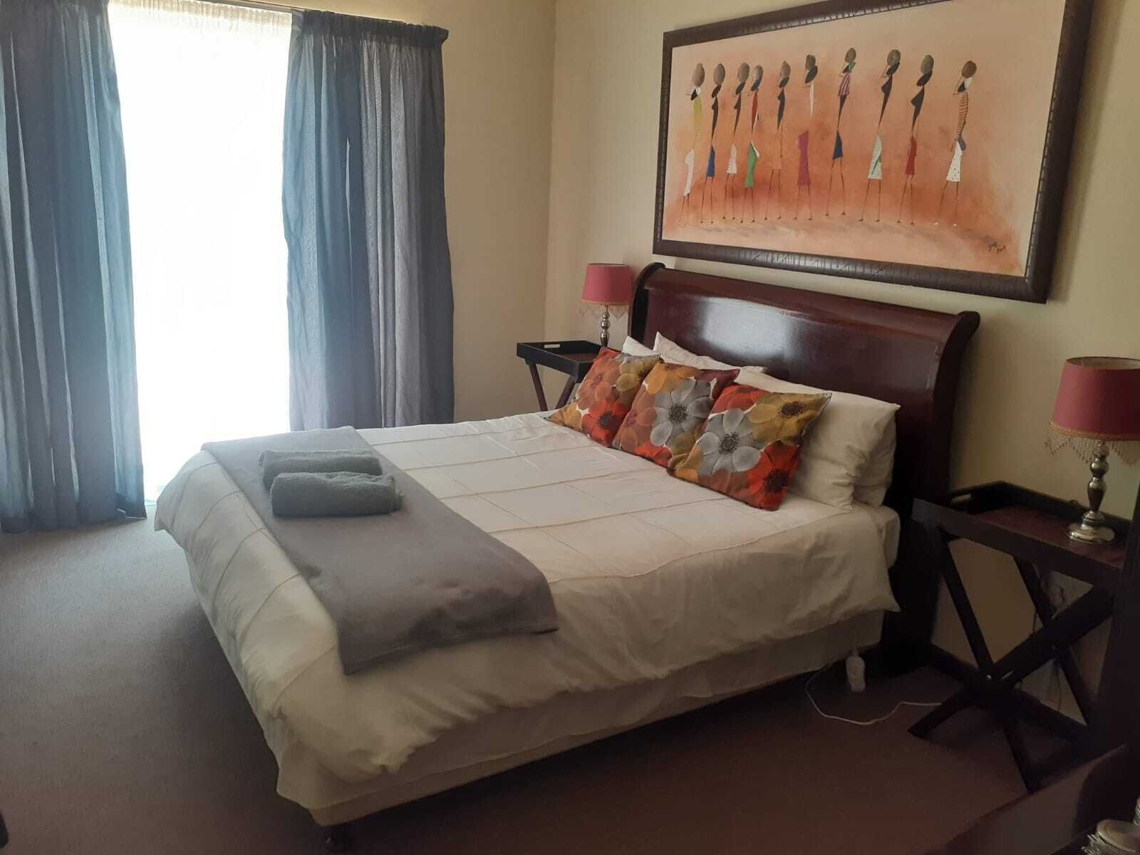 A hotel room with a bed, two bedside tables, a lamp, a painting on the wall, and a window with curtains.