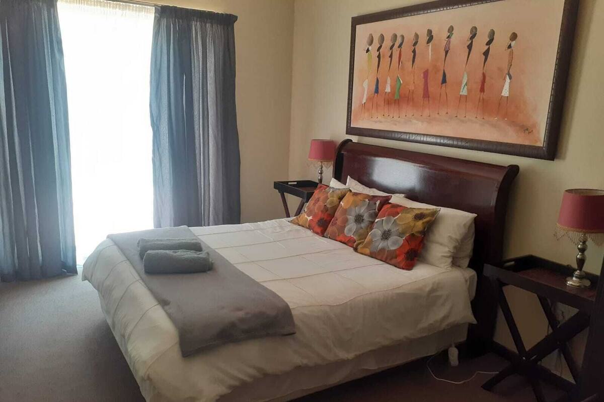 Habitación de hotel con cama, dos mesitas de noche, lámpara, un cuadro en la pared y una ventana con cortinas.