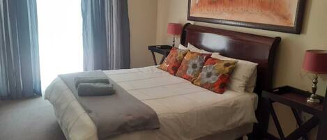 Habitación de hotel con cama, dos mesitas de noche, lámpara, un cuadro en la pared y una ventana con cortinas.