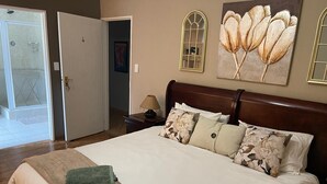 Un dormitorio con cama, almohadas, una mesita de noche, lámpara y puerta que da a un baño.