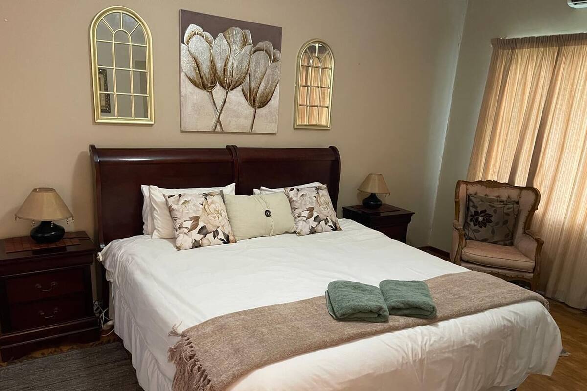 Una cama bien hecha con cabecera marrón, dos mesitas de noche con lámparas, un cuadro floral sobre la cama y una silla con cojín floral.