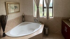 A bathroom with a large bathtub, a window with curtains, a framed picture on the wall, and a trash can.