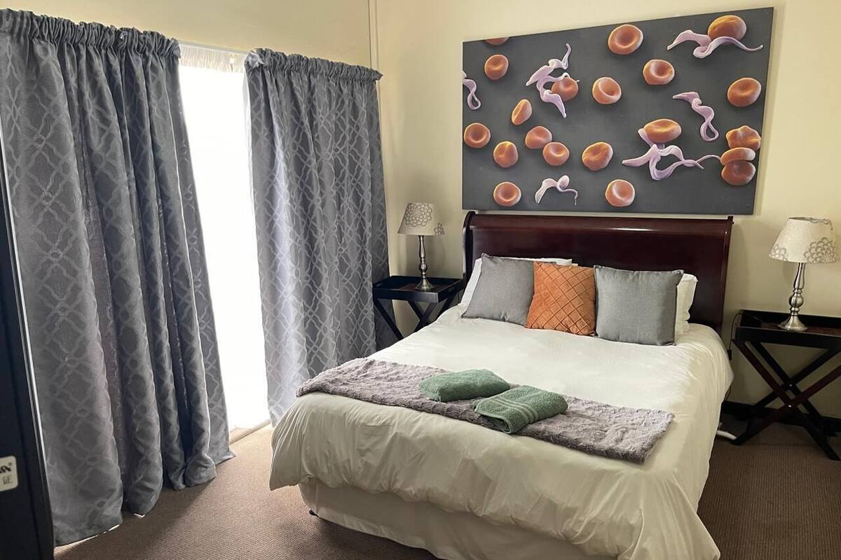 Habitación de hotel con cama, dos mesitas de noche, una lámpara, un cuadro y cortinas.