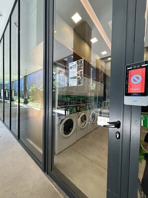 A public laundromat with industrial-style washers and dryers, a glass wall, and a digital payment kiosk. - Millenium Luxury | Cozy Studio in Vila Olímpia (São Paulo)