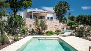 Ein modernes Haus mit Steinaußenwand, einem Schwimmbad und einem Garten mit verschiedenen Pflanzen und einer Bank.