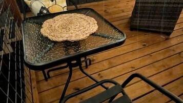 A wooden deck with a metal table and chairs, a woven mat on the table, and a yellow pillow with text.