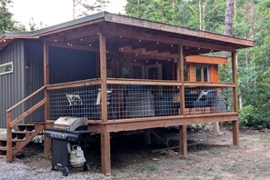 Een houten hut met een overdekte veranda, een barbecue en een vuilnisbak.