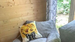 A corner of a room with a wooden wall, a window with a floral curtain, and a grey upholstered chair with a sunflower pillow. - Luxury Woodland Cabin Near The Caverns & Savage Gulf, Tennessee (Coalmont)