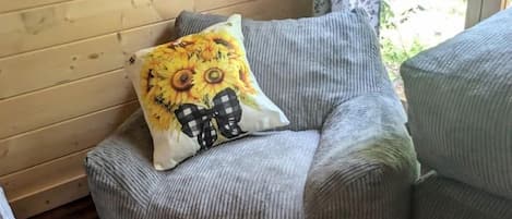 Esquina de una habitación con pared de madera, una ventana con cortina floral y un sillón tapizado en gris con un cojín de girasol.