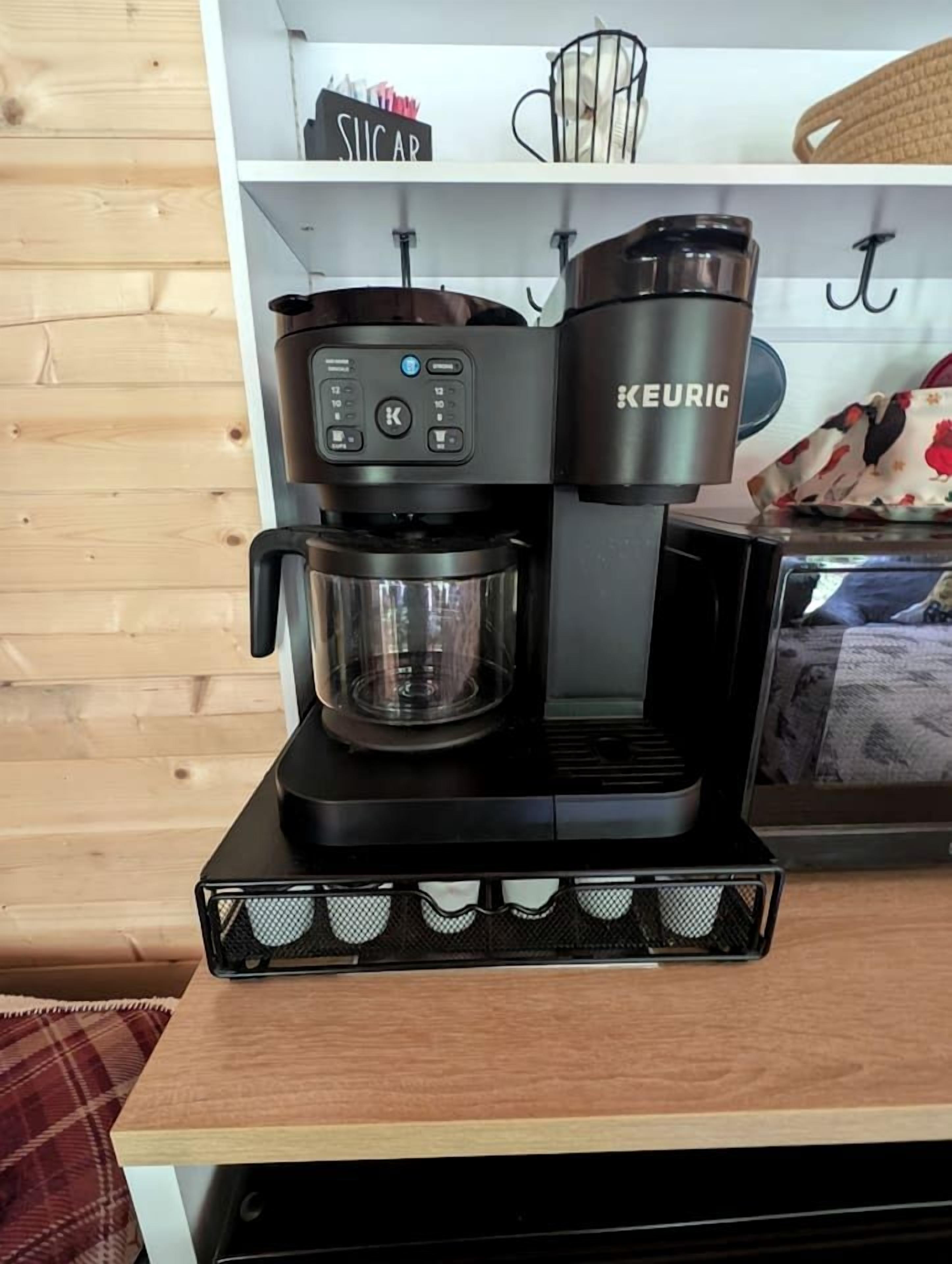 A black Keurig coffee maker on a wooden table with a coffee pot and a mug.