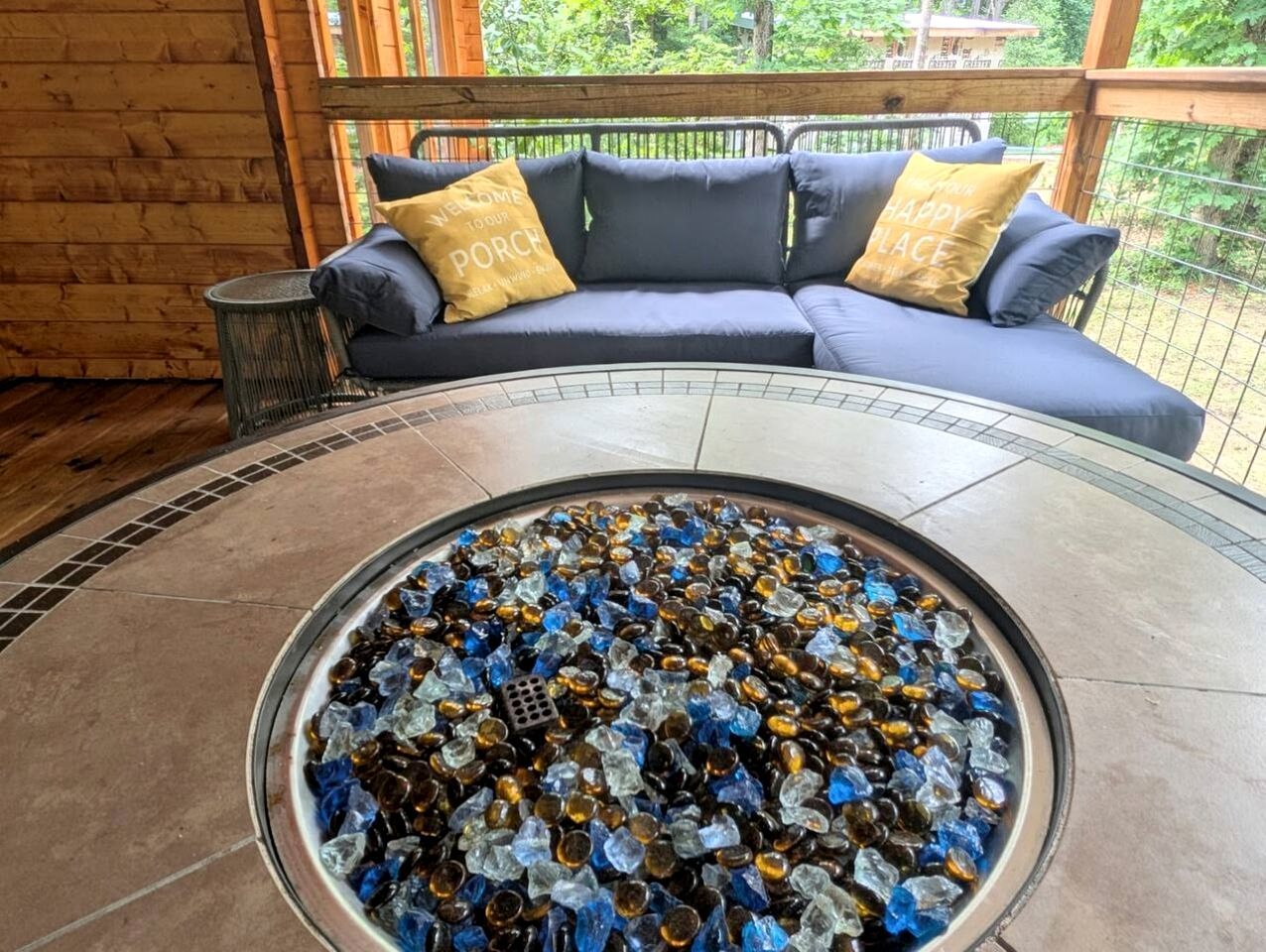Una mesa redonda de vidrio con un mosaico de piezas de vidrio, un sofá azul con cojines amarillos y un interior de madera.