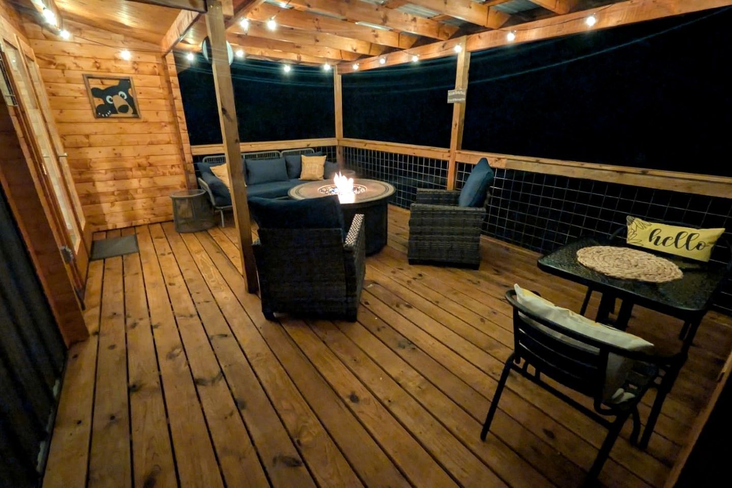 Eine Holzterrasse mit einem runden Tisch, zwei Stühlen, einem kleinen Tisch mit einem 'hello' Schild und einer Feuerstelle.