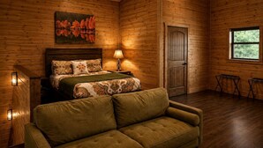 A log cabin bedroom with a bed, a sofa, a painting, a window, and a door. - Luxury Trailside Cabin with Hot Tub near Glover River in Broken Bow, Oklahoma (Broken Bow)
