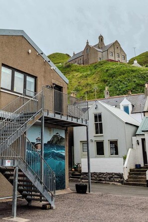 Un edificio costero con un mural, una escalera y vista a una casa en la colina.