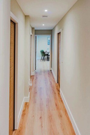 Un pasillo con suelo de madera, paredes blancas y un comedor visible en la distancia.