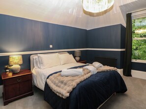 A bedroom with a bed, nightstands, a lamp, and a view of greenery through a window. - The Loft Suite (Buxton)