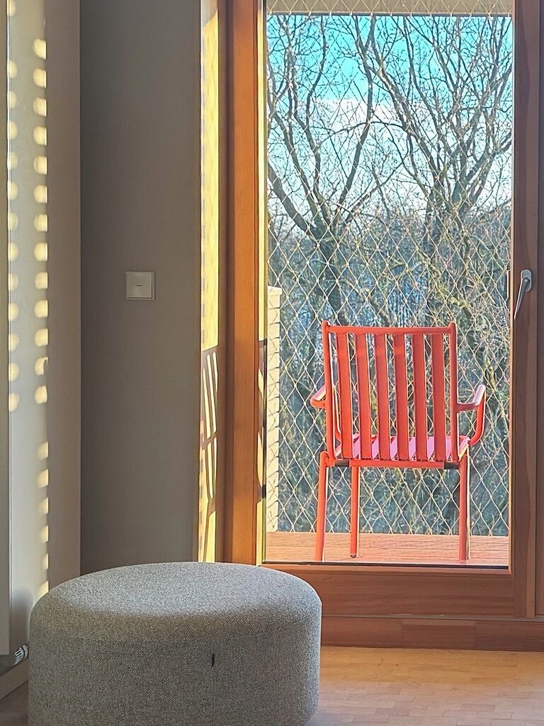 A room with a wooden bench, a grey ottoman, and a view through a glass door to an outdoor area with trees.