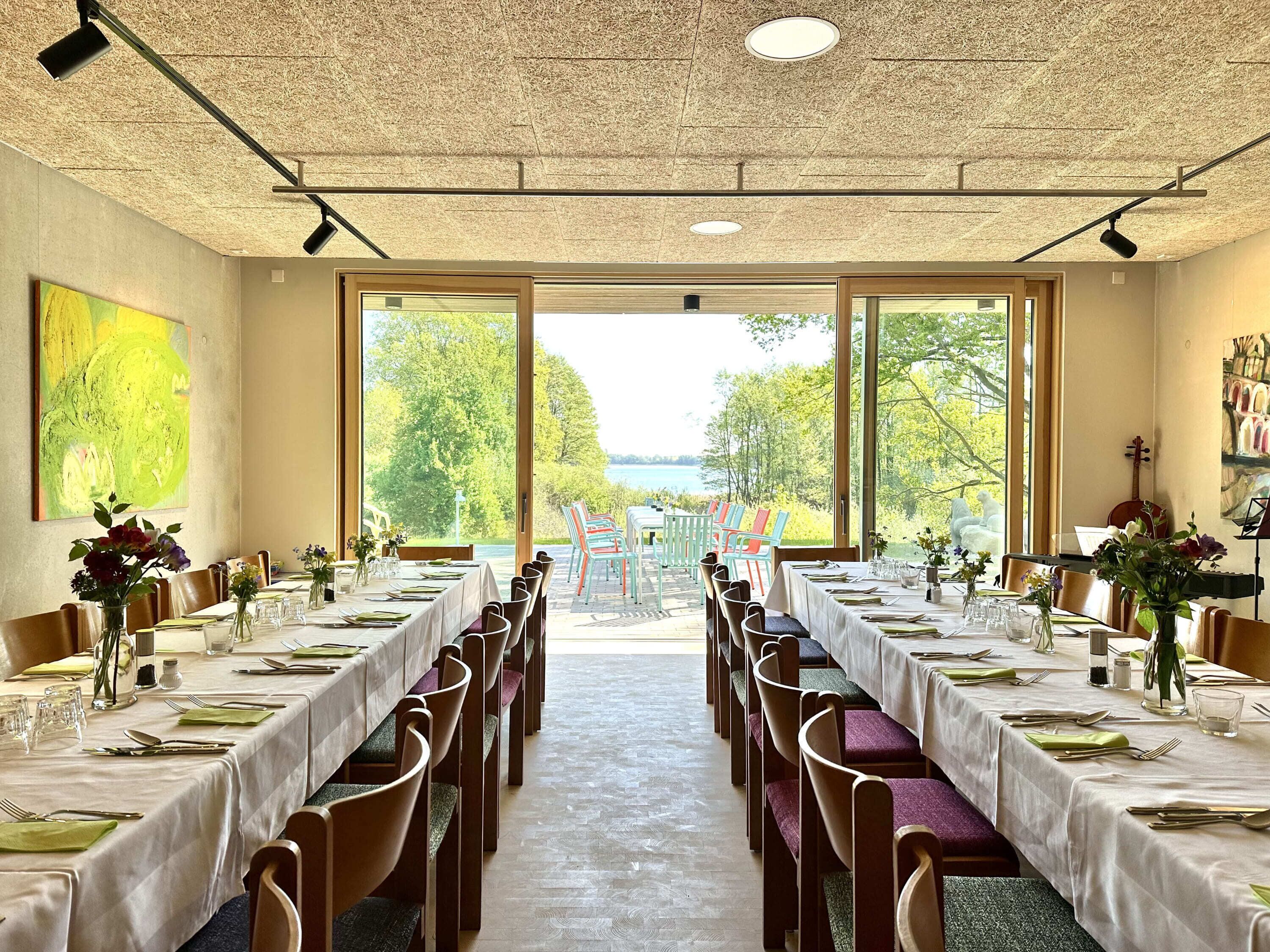 A dining area with tables set for service, large windows with a view of trees and a body of water, and a ceiling with a textured pattern.