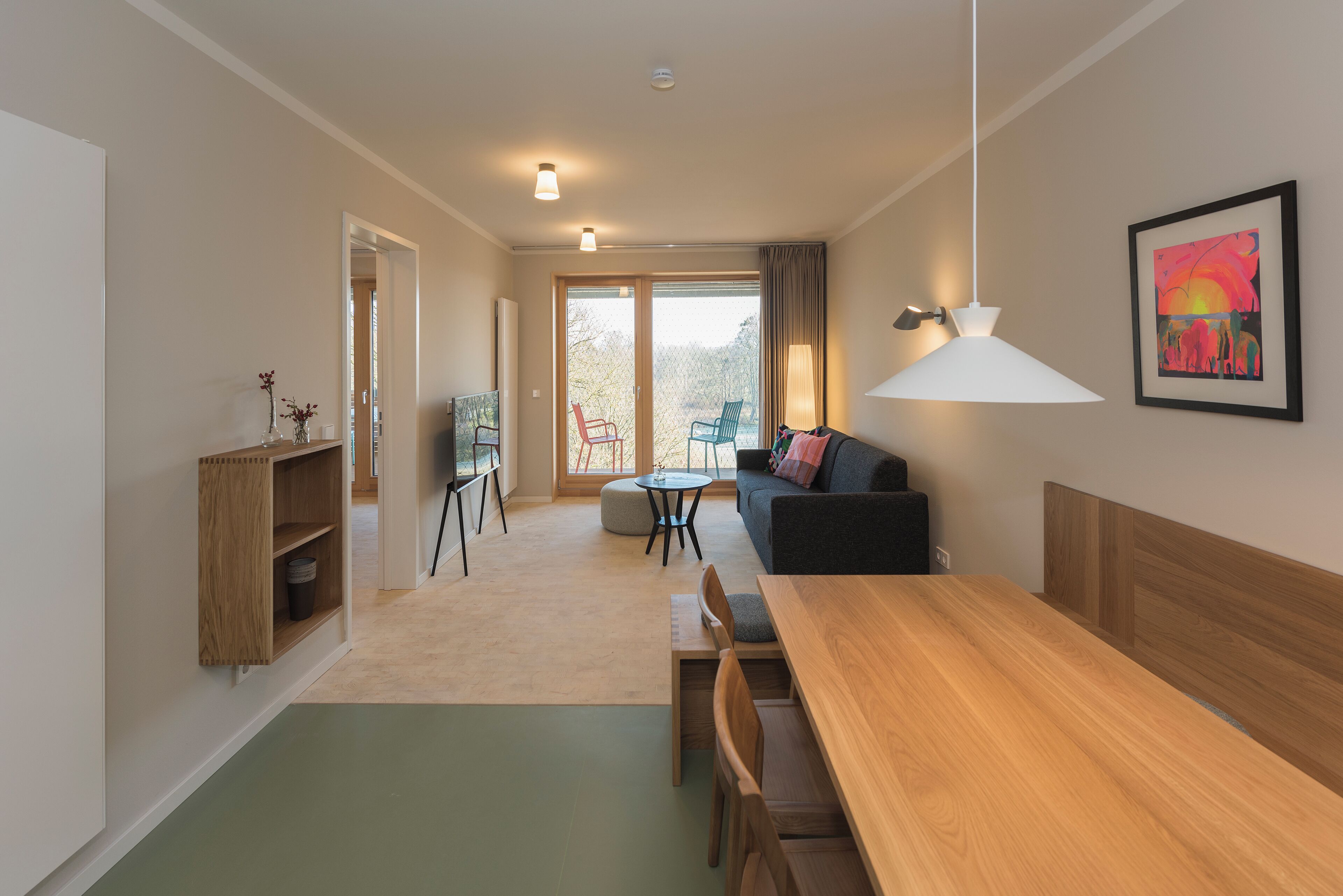 Ein modernes Wohnzimmer mit einer Couch, einem Esstisch und Stühlen. Es gibt einen Holzkasten, eine Lampe und ein Bild an der Wand.
