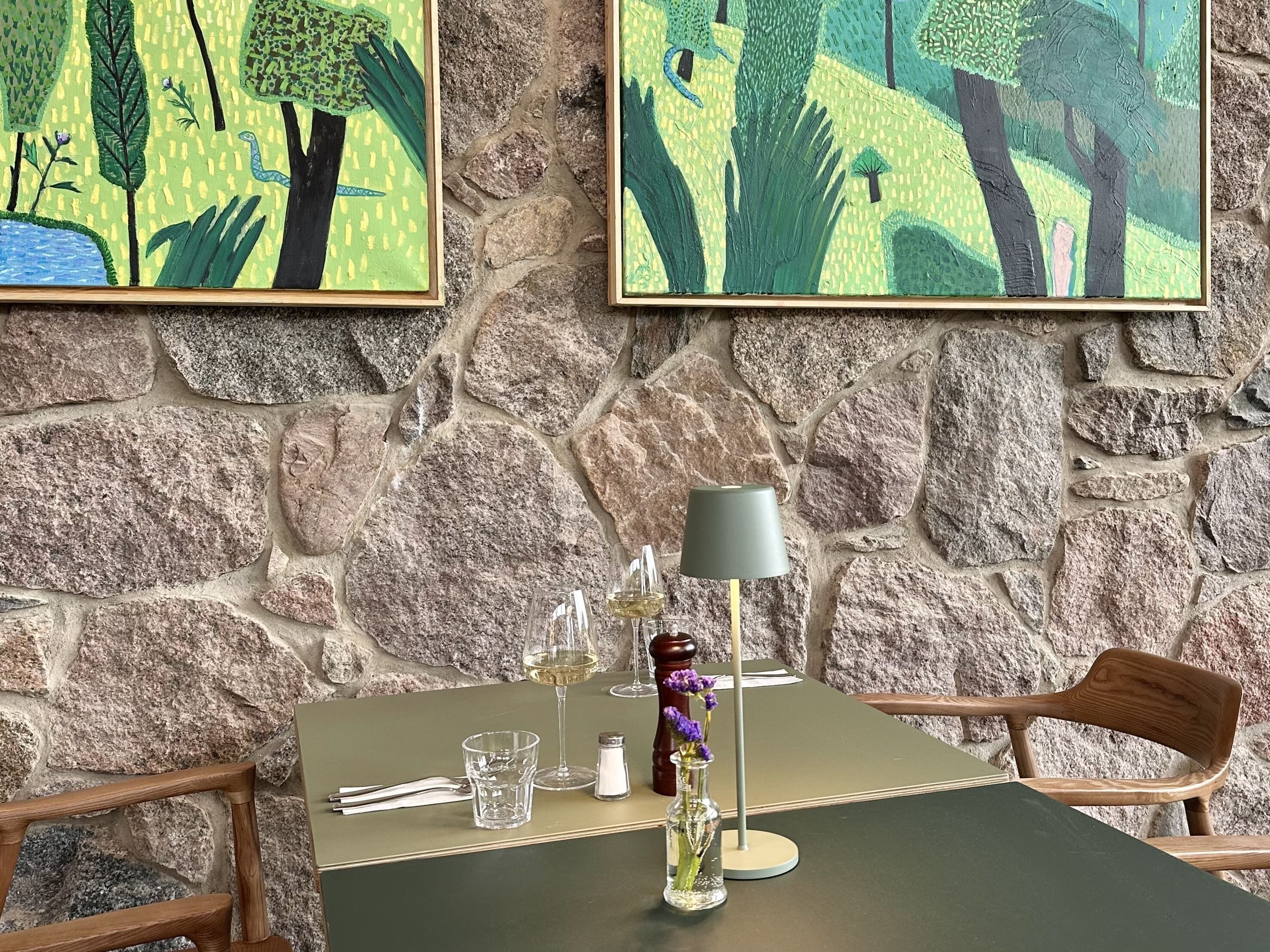 Eine Steinsäule mit zwei gerahmten Gemälden, ein Tisch mit Lampe, Weinglas und einer Blumenvase sowie Holzstühle.