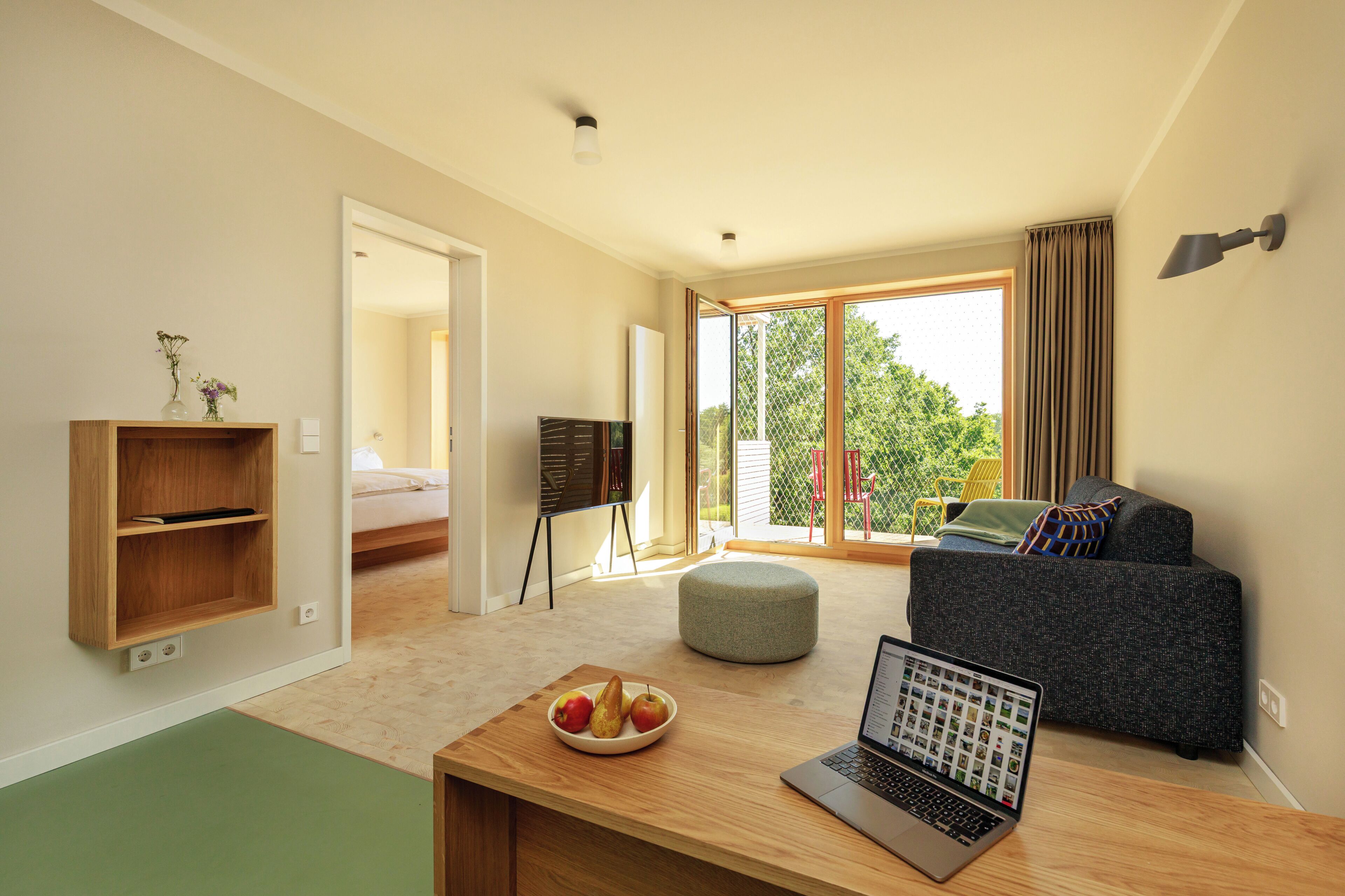 A modern living room with a laptop, a bowl of fruit, a grey ottoman, a grey sofa, and a wooden shelf.