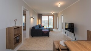 A modern living room with a sofa, a wooden table, a television, and a view through a large window. - SEEZIEGE Ferienwohungen in der Uckermark (Boitzenburger Land)