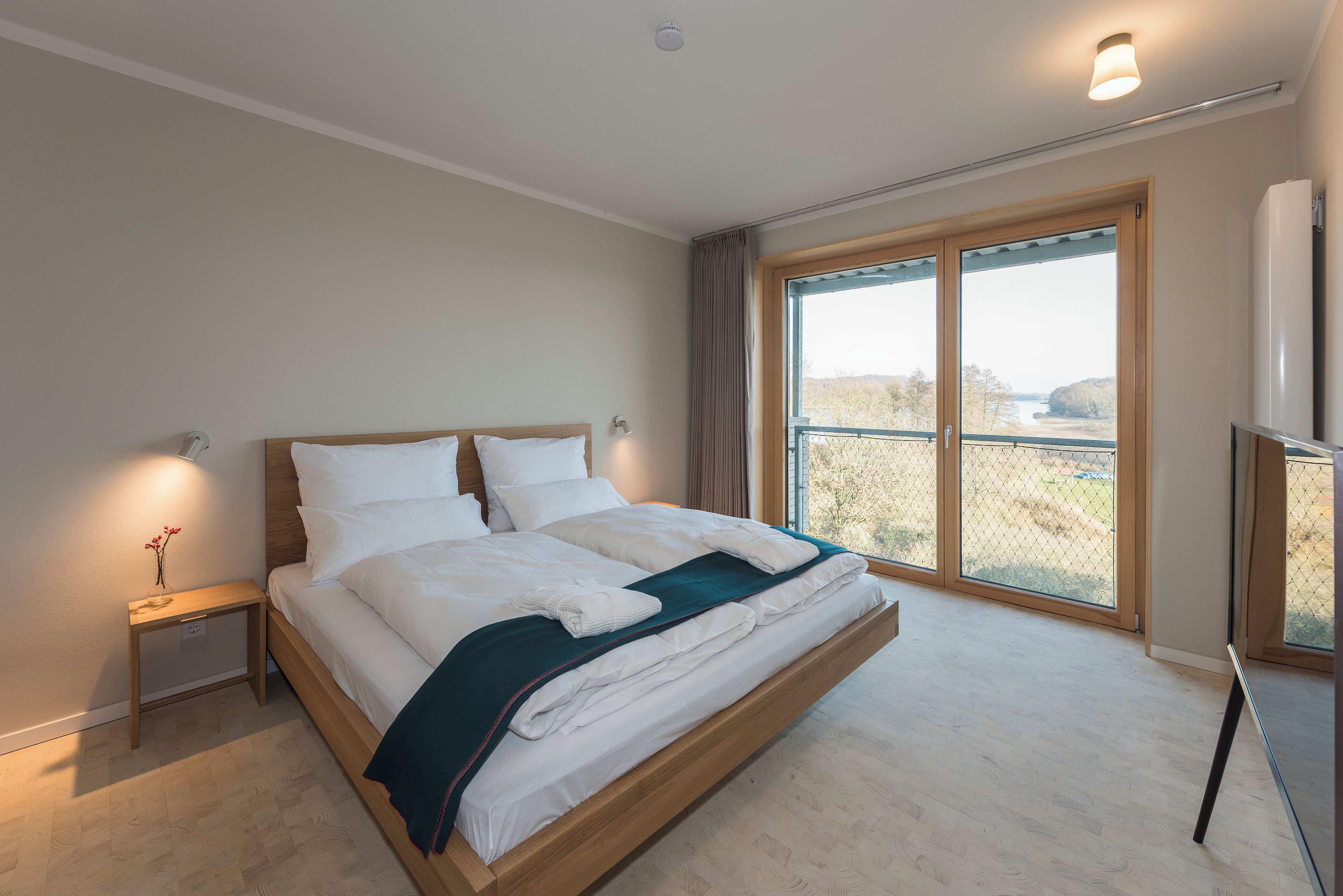 A bedroom with a large bed, a nightstand, a lamp, a window with a view, and a refrigerator.