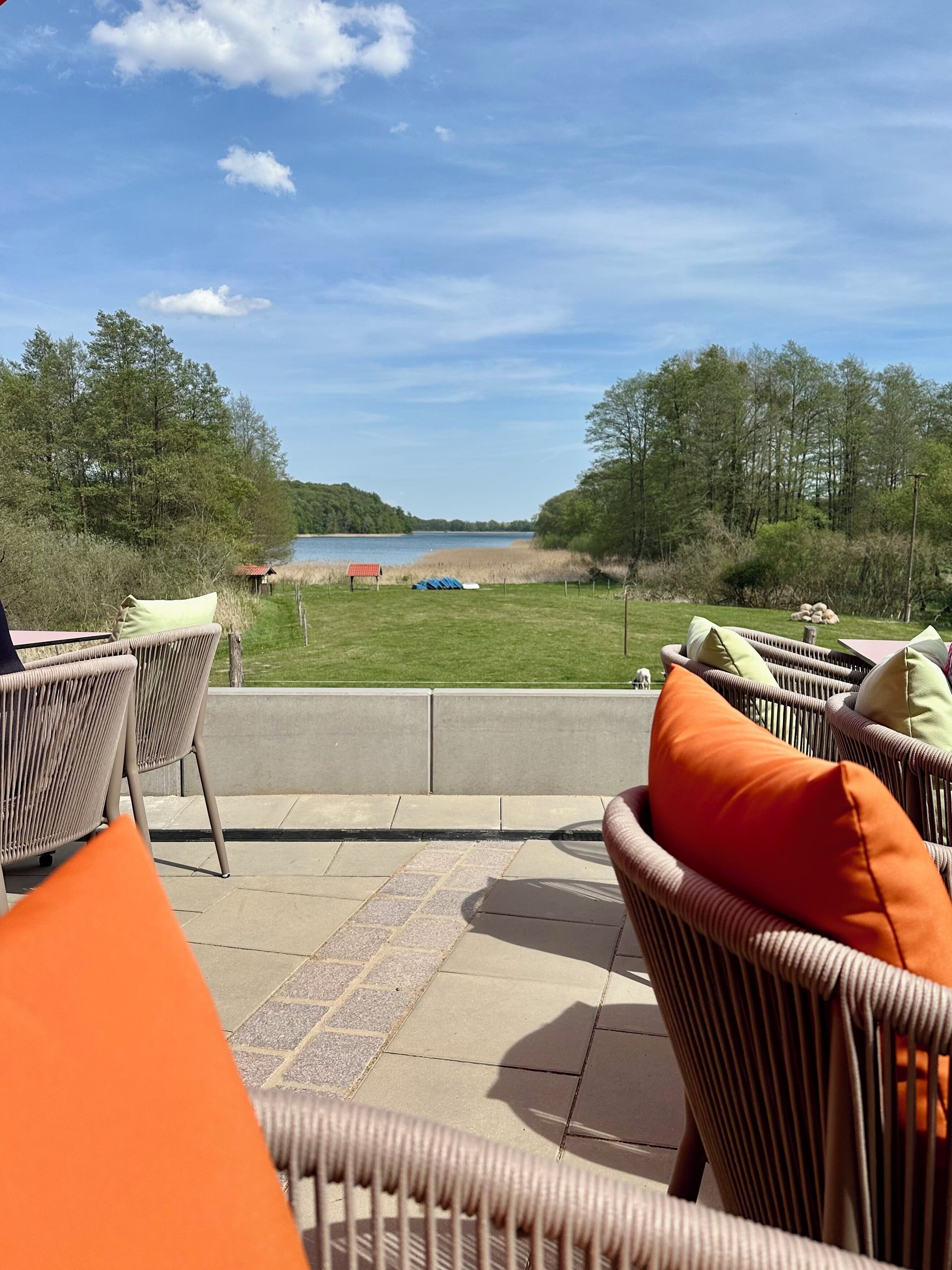Ein Sitzbereich im Freien mit Korbstühlen und orangefarbenen Kissen mit Blick auf einen See und eine Rasenfläche.