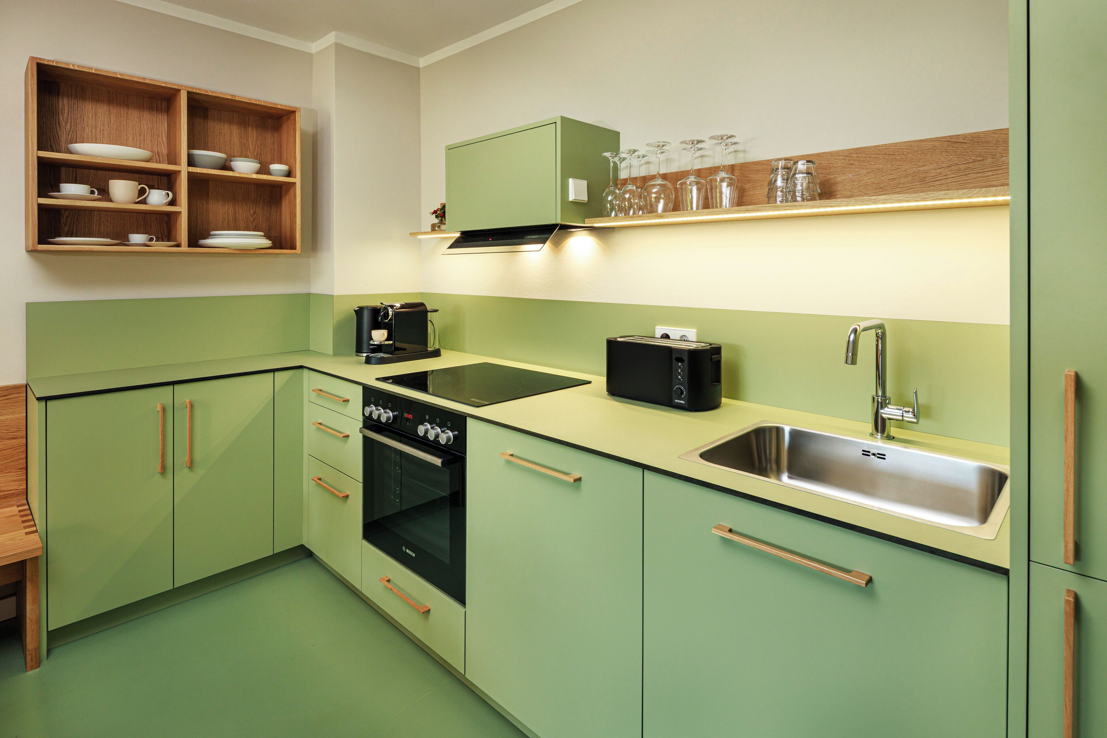 A kitchen with green cabinets, a black stove, a sink, and a shelf with glassware.