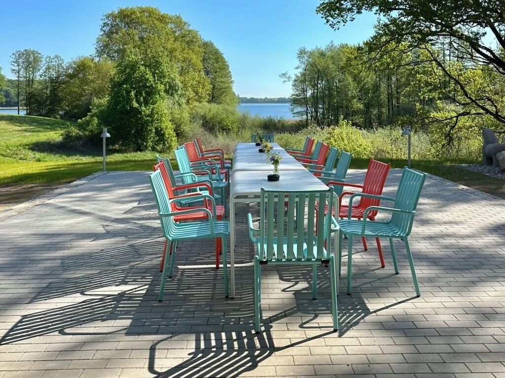 Un área de comedor al aire libre con una larga mesa y sillas, rodeada de árboles y vistas a un cuerpo de agua.