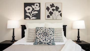 Ein ordentlich bezogenes Bett mit einem gemusterten Kissen, zwei Nachttischlampen und zwei gerahmte Blumenbilder an der Wand.