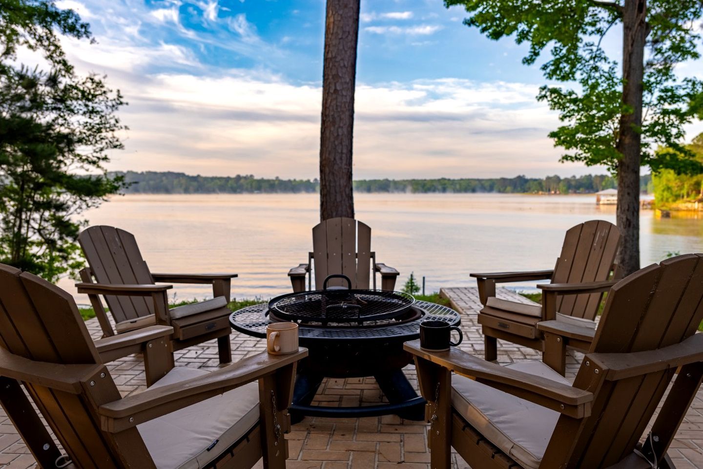 A patio with Adirondack chairs, a round table, and a fire pit overlooking a lake.