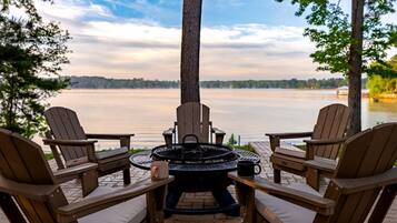 A patio with Adirondack chairs, a round table, and a fire pit overlooking a lake.