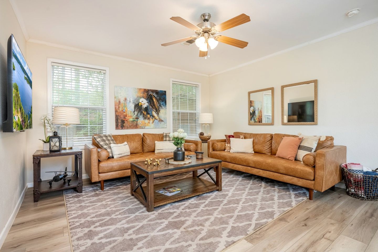A living room with a brown leather sofa, a wooden coffee table, and a ceiling fan. There are two windows with blinds, a TV mounted on the wall, and a small side table with a lamp.