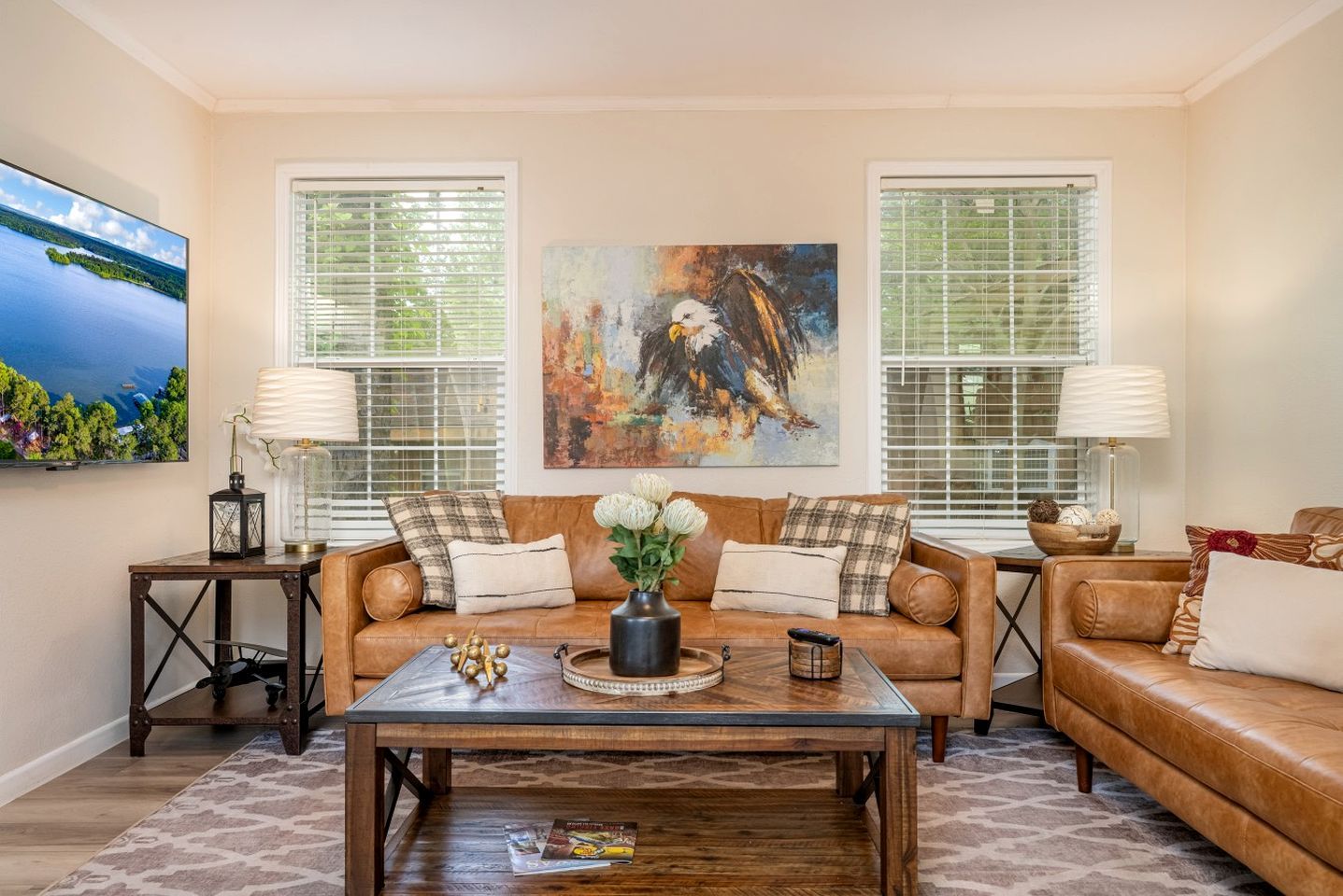 A living room with a brown leather sofa, a coffee table, and two side tables with lamps. There are two windows with blinds, a painting on the wall, and a television on the left.