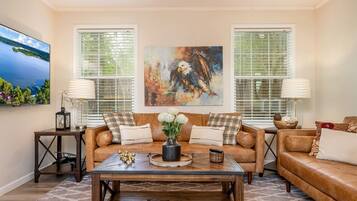 A living room with a brown leather sofa, a coffee table, and two side tables with lamps. There are two windows with blinds, a painting on the wall, and a television on the left.