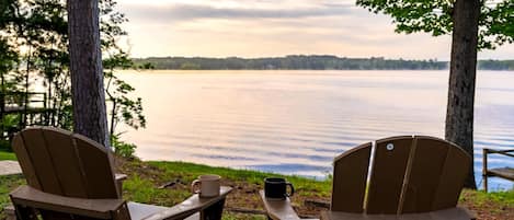 Dos sillas Adirondack mirando un lago tranquilo con una orilla arbolada y un cielo nublado.