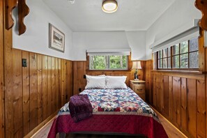 Ein Schlafzimmer mit Holzvertäfelung, einem Bett mit Steppdecke, einem Fenster mit Blick auf Bäume und einem Nachttisch mit Lampe.