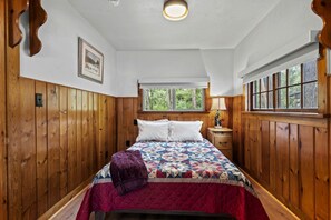 A bedroom with wooden paneling, a bed with a quilt, a window with a view of trees, and a nightstand with a lamp. - Charming Pet-Friendly Cabins with Scenic Outdoor Spaces near Garden of the Gods in Colorado Springs, Colorado (Colorado Springs)