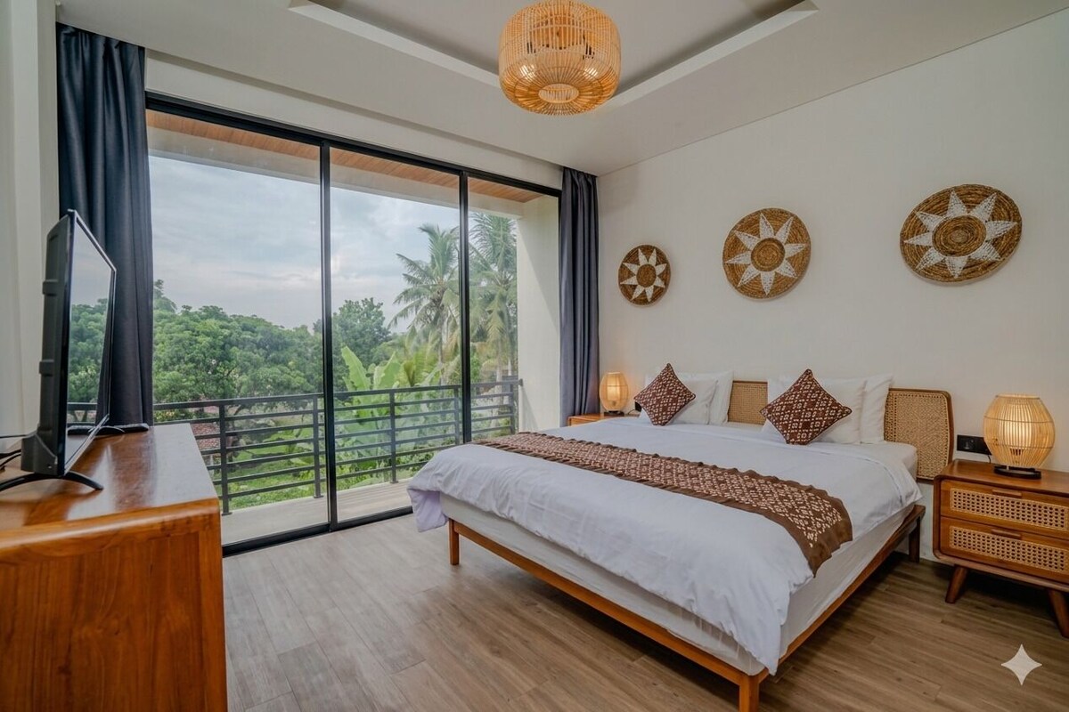 A bedroom with a large bed, a desk, a TV, and a balcony with a view of greenery.