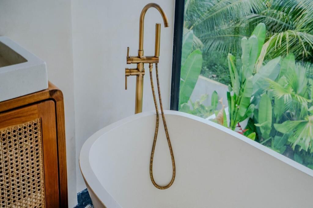 A bathroom with a freestanding bathtub, a brass faucet, and a view of lush greenery through a window.